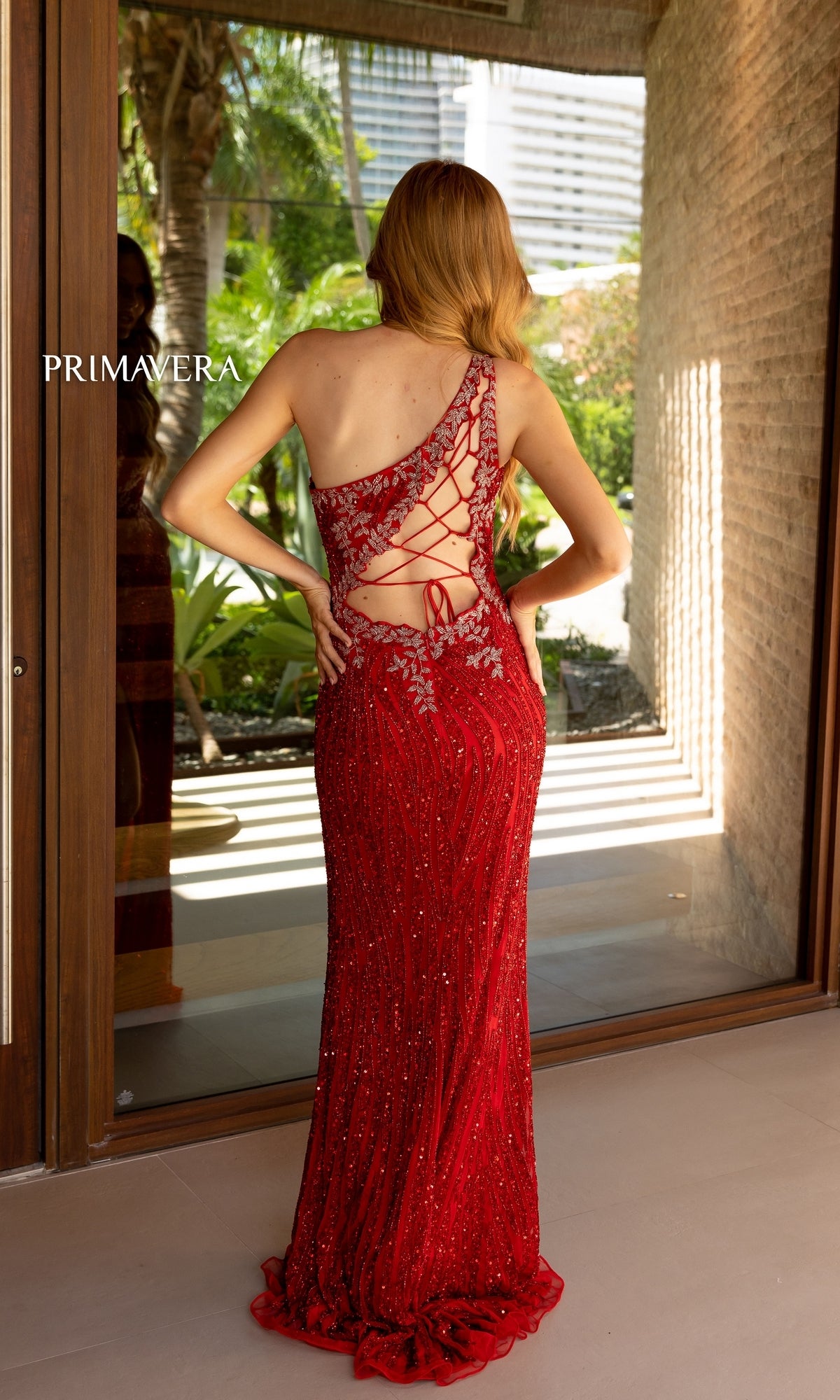 Wearing the Primavera One-Shoulder Beaded Long Prom Dress 4144, a woman stands with her back to a glass door, framed by tropical plants and buildings in the background.